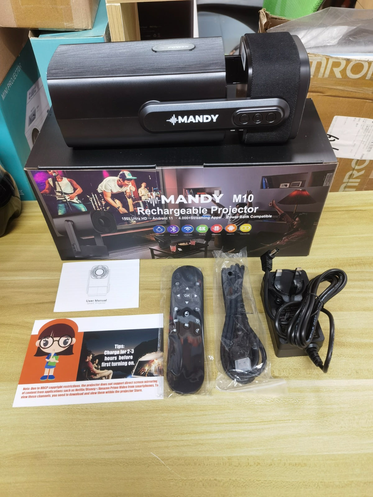 Mandy M10 rechargeable projector shown with its retail box, remote control, power cable, and instruction booklet on a light wood tabletop.