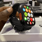 Mandy Android Smartwatch M12 with colorful app icons held by a hand on a desk with a laptop and keyboard in the background.